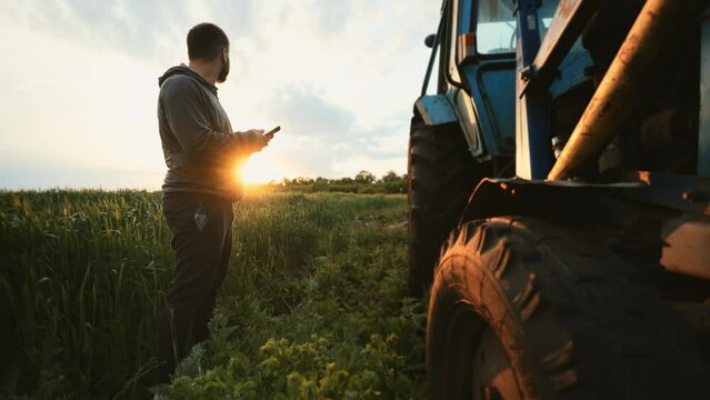 Farmer man stands in green field near tractor and uses mobile phone for text messaging or remote control of agriculture. Break rest in working day. Wireless technology. Communicate on social networks.