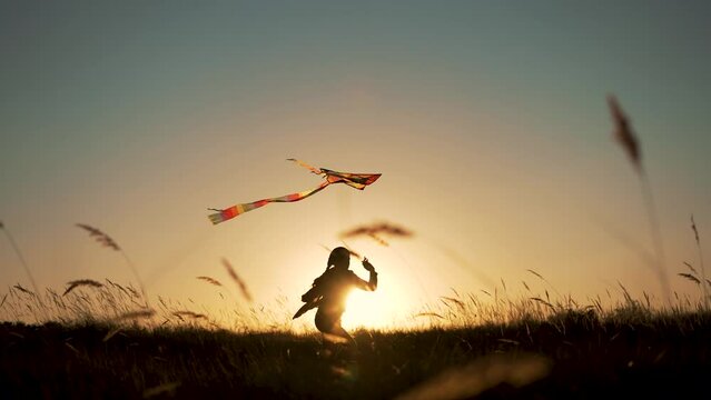 Happy child. Girl with flying kite in park. Child runs at sunset on grass in park. Silhouette of girl with flying kite. Child launches flying kite into sky in the wind. Silhouette of child in meadow
