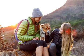 Smiling young woman in down jacket sitting with friend on rock with their pet dog