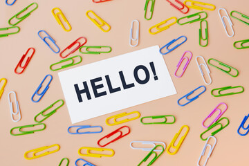 Hello salutation or greeting word to welcome someone or initiate a conversation. Communication concept, introduction. Top view of business card and many colourful paper clips on beige background