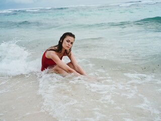 A woman in a red swimsuit sits on the sand by the ocean in the waves on the beach of Bali Island and smiles