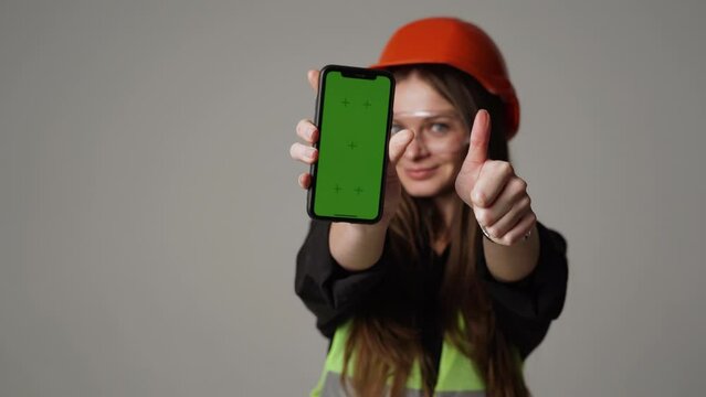 Pretty woman engineer in orange helmet and goggles holding green screen phone.Construction and industrial design concept