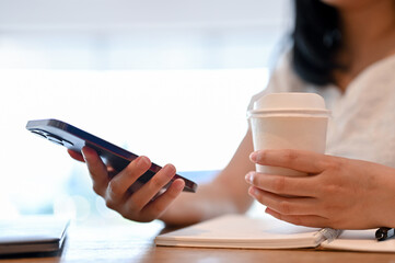 Asian woman using her smartphone and sipping coffee while relaxing in the coffee shop. cropped