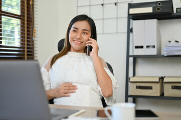 Successful Asian pregnant female boss talking on the phone while touching her belly at her desk.