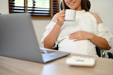 Asian pregnant female boss or businesswoman sipping tea while working in her office. cropped