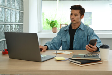 Handsome hipster man using laptop computer and smart phone at his workstation