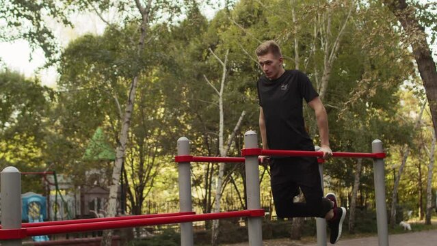 Young caucasian sportsman doing dips on an outdoor workout area. Athlete with short haircut in black t-shirt doing exercises on a parallel bars