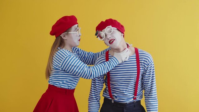 Young pantomime woman strangling and emotionally yelling at young pantomime man as a joke. Two mimes starting funny performance on yellow background
