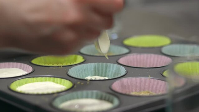 A woman scooping cheesecake cream filling with a spoon to fill mini muffin holders, which sit in a muffin pan, to make cheesecake cups. the muffin holders are blue, pink and yellow.