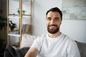 cheerful and bearded man in white t-shirt sitting on couch in modern living room.