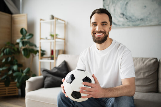 bearded sports fan sitting on couch in blurred living room and smiling at camera.