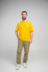 full length of happy bearded man in beige pants and yellow t-shirt standing with hand in pocket on grey background.