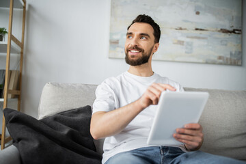 happy man in white t-shirt sitting on couch and using digital tablet in modern living room.