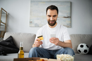 KYIV, UKRAINE - OCTOBER 21, 2022: cheerful man holding credit card and using smartphone while doing online shopping.