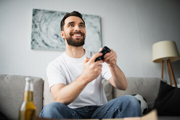 KYIV, UKRAINE - OCTOBER 21, 2022: happy bearded man playing video game near bottle of beer at home.