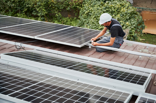Man technician mounting photovoltaic solar moduls on roof of house. Builder in helmet installing solar panel system outdoors. Concept of alternative and renewable energy.
