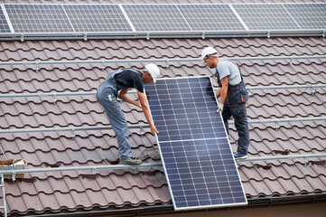 Men technicians lifting up photovoltaic solar moduls on roof of house. Engineers in helmets...
