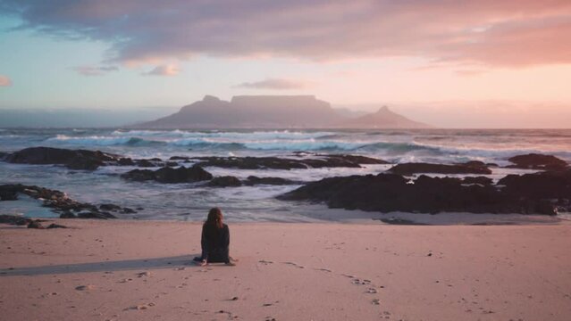 Woman is sitting on the beach in a summer dress looking at the sea and enjoying the view, Cape Town.