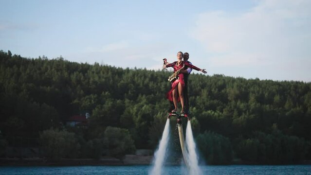 Show over the water on a flyboard. A woman in a red dress plays the saxophone, a man flies with her together.