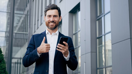 Happy man standing outdoors holding phone smiling businessman using new mobile app for business tasks showing thumb up gesture of approval demonstrating sign excellent result recommends best quality