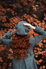 Young elegant girl or woman with beautiful long curvy red hair standing with her back, her hands on head in autumn park, orange leaves background, warm vintage outdoor seasonal lifestyle portrait