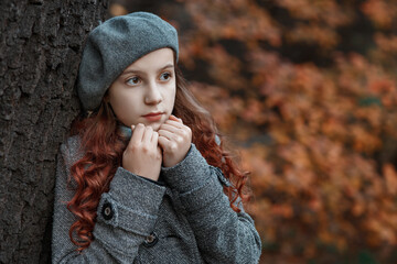 Young serious gloomy sad girl with long red hair in grey coat, beret  and scarf standing near tree...