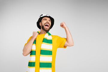 overjoyed man in striped scarf and football fan hat screaming and showing triumph gesture isolated on grey.