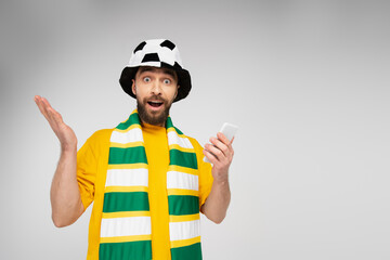 surprised man in striped scarf and football fan hat holding cellphone and looking at camera isolated on grey.