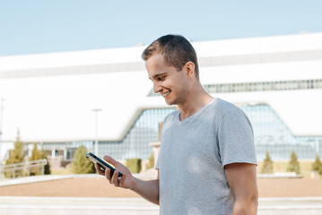 Smiling young man browsing internet on phone outdoors. Side view portrait of guy having fun while looking into smartphone screen