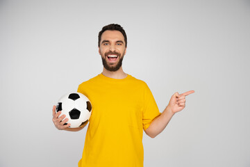 football fan laughing at camera and pointing with finger while holding soccer ball isolated on grey.