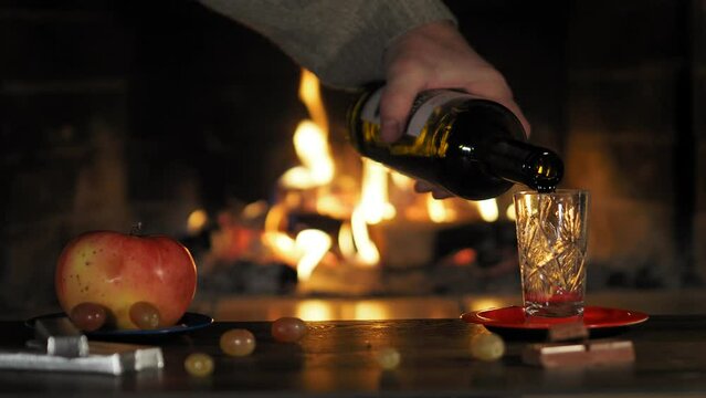 Drinks. A man pours a drink from a bottle into a glass standing on the table next to an apple, grapes and chocolate slices against the background of a fireplace flame. Close-up.