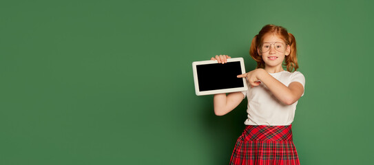 Happy charming girl, kid pointing at tablet screen. Student or pupil of elementary school wearing...