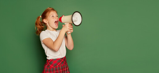 Back to school concept. Happy pupil of elementary school wearing modern school uniform isolated over dark green background. Happy charming girl, kid with megaphone