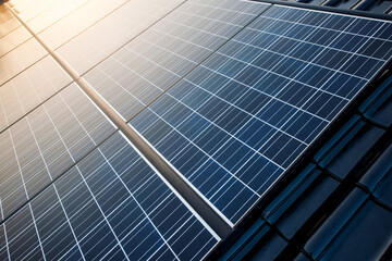 New solar panels are mounted on a roof with shiny black tiles and the clear sky is reflected in the warm sunlight in it.