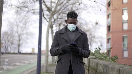 Black man wearing covid-19 face mask while walking outside looking at cellphone