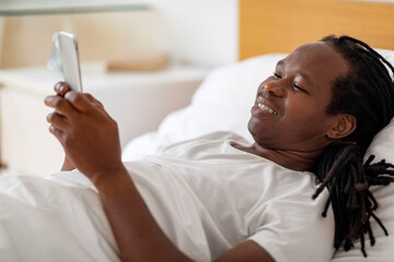 Gadget Addiction. Black Man Browsing Internet On Smartphone While Resting In Bed