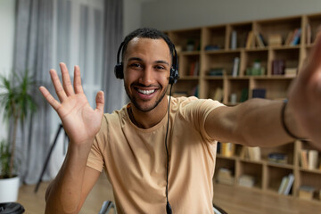 Fototapeta premium Online communication. Happy arab man in headphones making video call to friends or colleagues, waving hand at camera