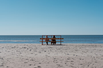 Dwie kobiety siedzą na ławce na piaszczystej plaży w słoneczny dzień - obrazy, fototapety, plakaty