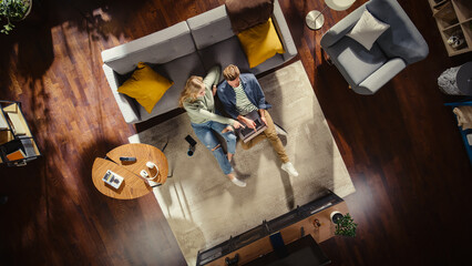 Top View Apartment: Happy Couple Using Laptop in the Stylish Living Room. Sitting on the Carpet and Browsing. On the Floor: Girlfriend and Boyfriend use Computer for Online Shopping, e-business.
