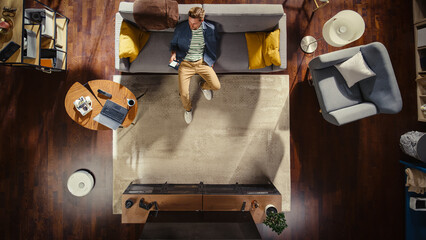 Apartment: Young Man Sitting on the Couch and Using Smartphone in Spacious Living Room. Guy Relaxes...