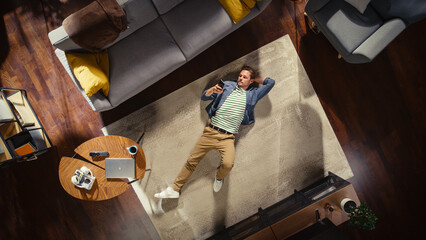 Fototapeta na wymiar Loft Apartment: Handsome Man Lying on a Living Room Floor, Using Smartphone. Guy Relaxes on the Carpet. Freelancer Does Remote Work, E-business, Online Shopping, Social Media Marketing. Top View.
