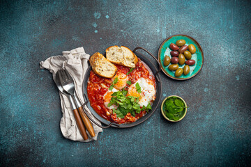 Middle Eastern and Maghrebi healthy dish Shakshouka made of eggs and tomato sauce served in pan with toasts, fresh cilantro and olives on rustic concrete background table from above