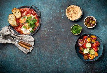 Middle Eastern traditional breakfast or brunch with eggs Shakshouka in pan with toasts, fresh vegetables salad, hummus and olives on rustic concrete background table from above, space for text