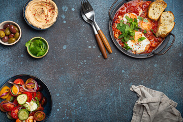 Middle Eastern traditional breakfast or brunch with eggs Shakshouka in pan with toasts, fresh vegetables salad, hummus and olives on rustic concrete background table from above, space for text