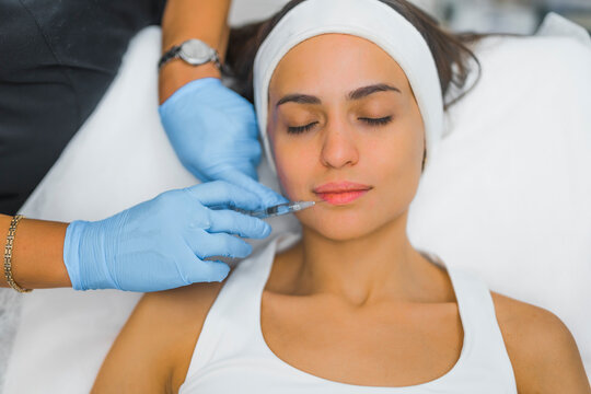 Medium closeup indoor view of relaxed caucasian woman in white tank-top during lip injection - lip filler - done by professional expert in protective gloves. High quality photo