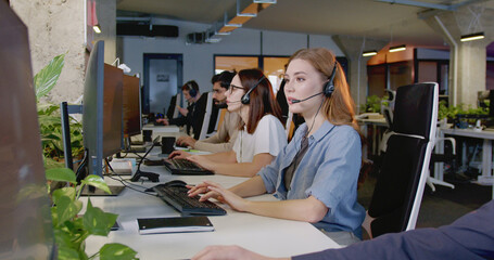 Caucasian beautiful cheerful woman in headset working at computer and talking with client in...