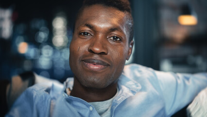 Obraz premium Close-up Face Portrait of the Young Handsome Black Man Smiling and Laughing at Something Funny on TV while Sitting on a Sofa in the Evening. Positive Human Emotions in Living Room Concept.
