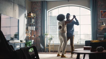Happy Black Couple Dances in the Living Room. Cheerful Girlfriend and Boyfriend Enjoy True Love, Good Mood and Energetic Music. Having Fun at Home. Stylish Interior and Sunny Day.