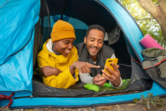 Happy Black Couple Using Cellphone And Sitting In Tent During Camping Having Fun Together