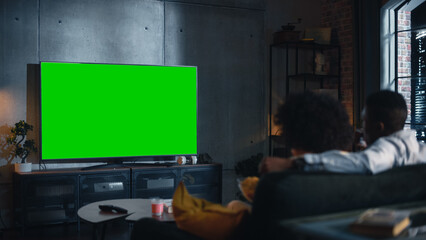 African American Couple Watching TV with Green Screen Mockup Display. They are Sitting on a Couch...